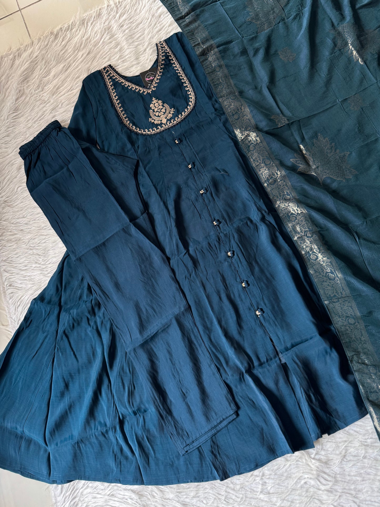 MD-201 Elegant deep teal ethnic ensemble featuring a flared kurta with delicate mirror buttons and intricate golden embroidery on the neckline, paired with matching pants and a rich, woven dupatta adorned with subtle floral motifs. Perfect for festive occ